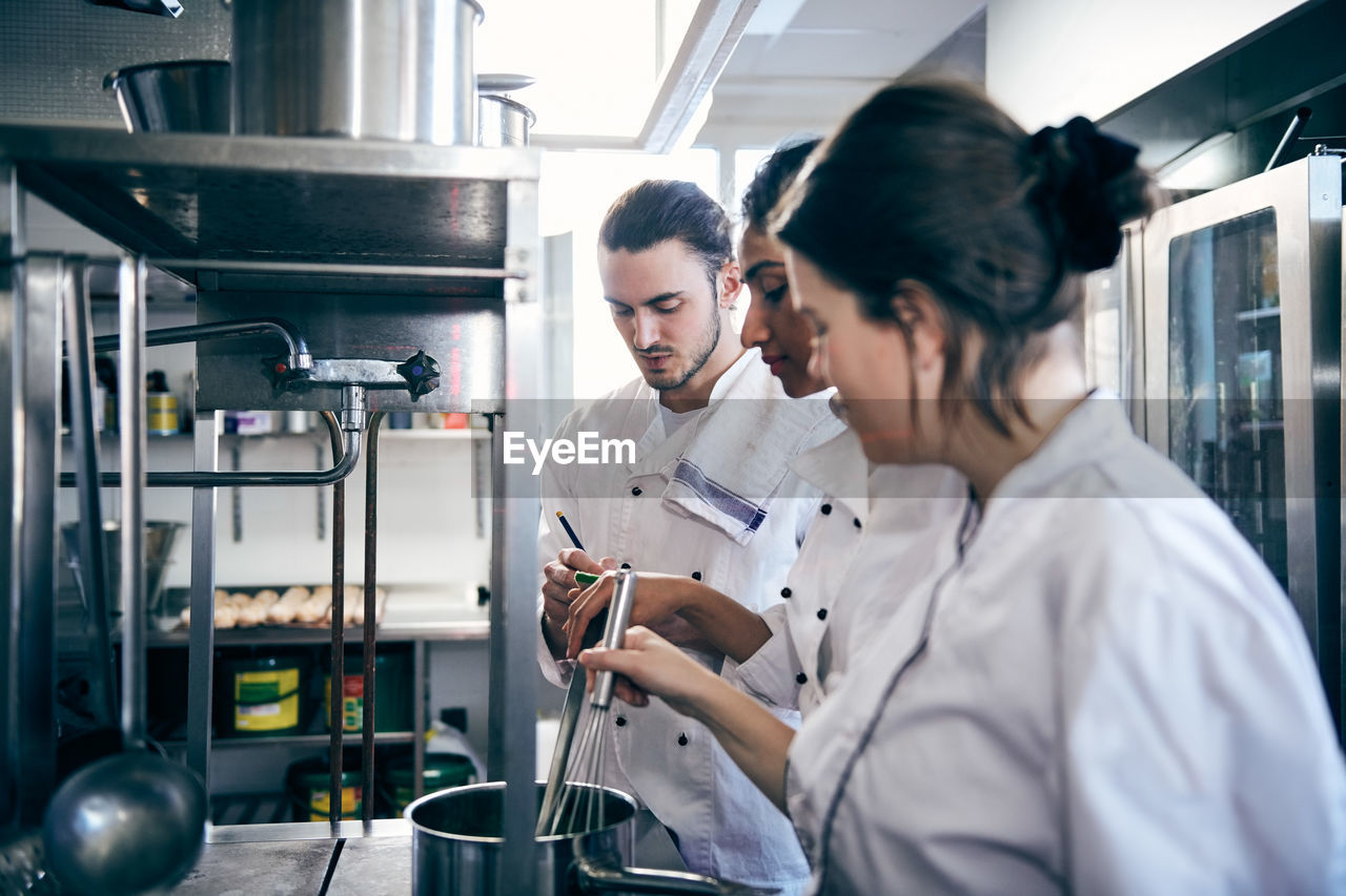 Female chefs cooking food while male colleague writing in notepad at commercial kitchen