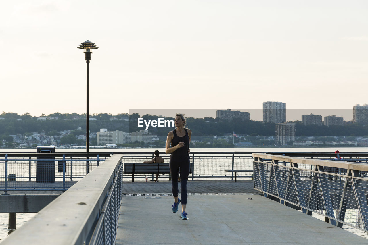 Woman jogging while exercising on bridge against clear sky in city