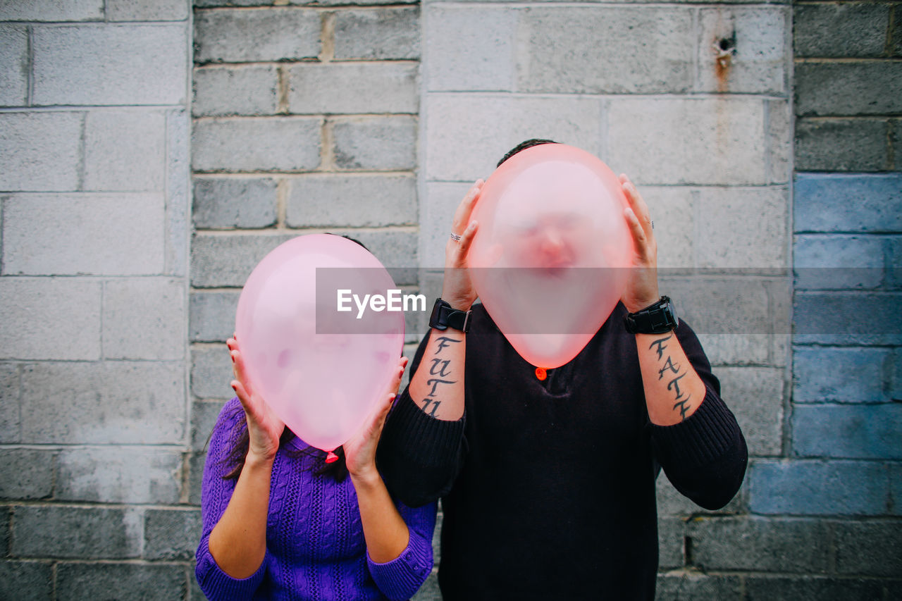 Close-up of man and woman holding balloons over face while standing against wall