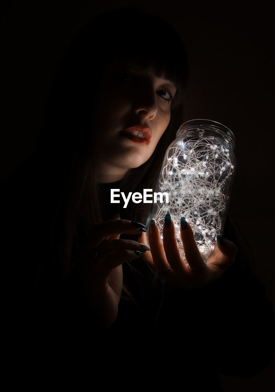 Portrait of young woman holding jar with illuminated string light against black background