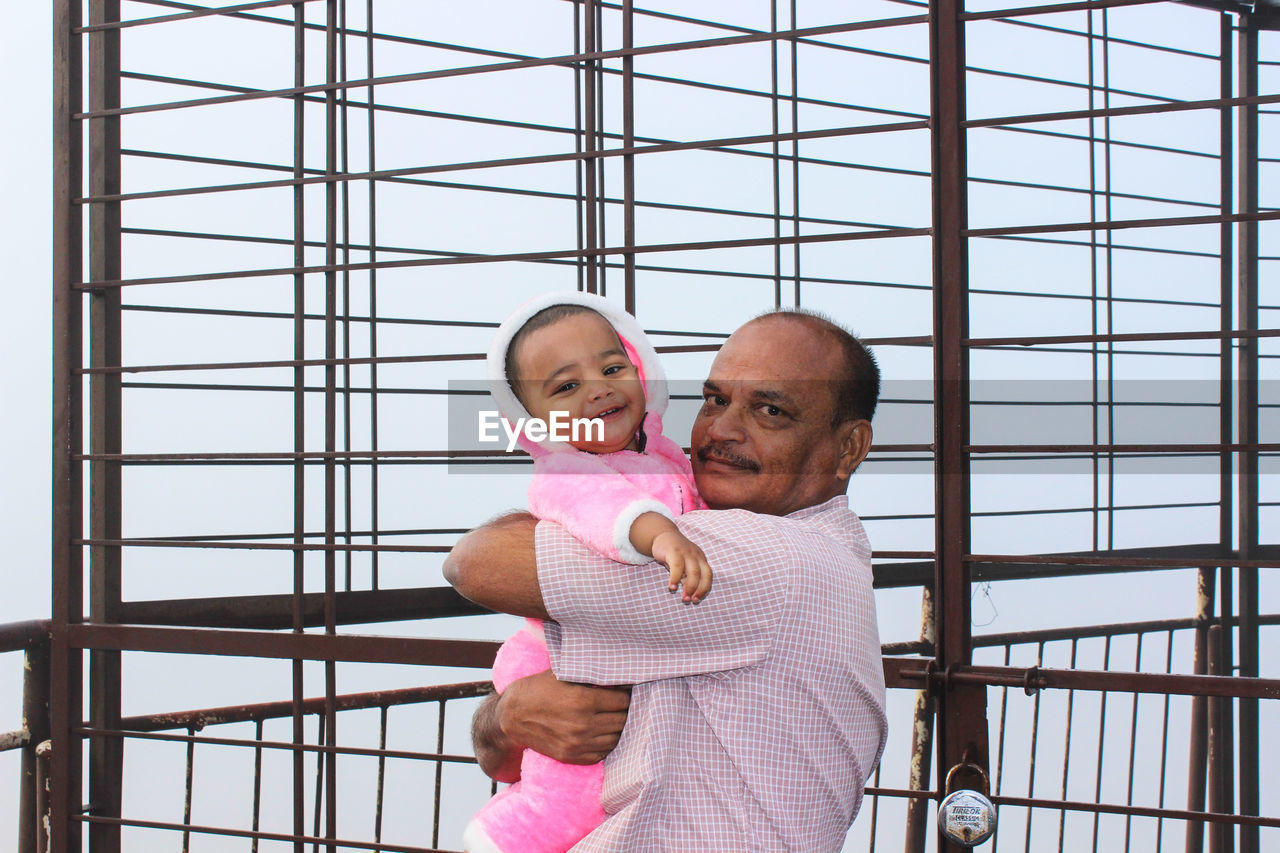 Side view portrait of grandfather carrying grandson while standing by metallic gate