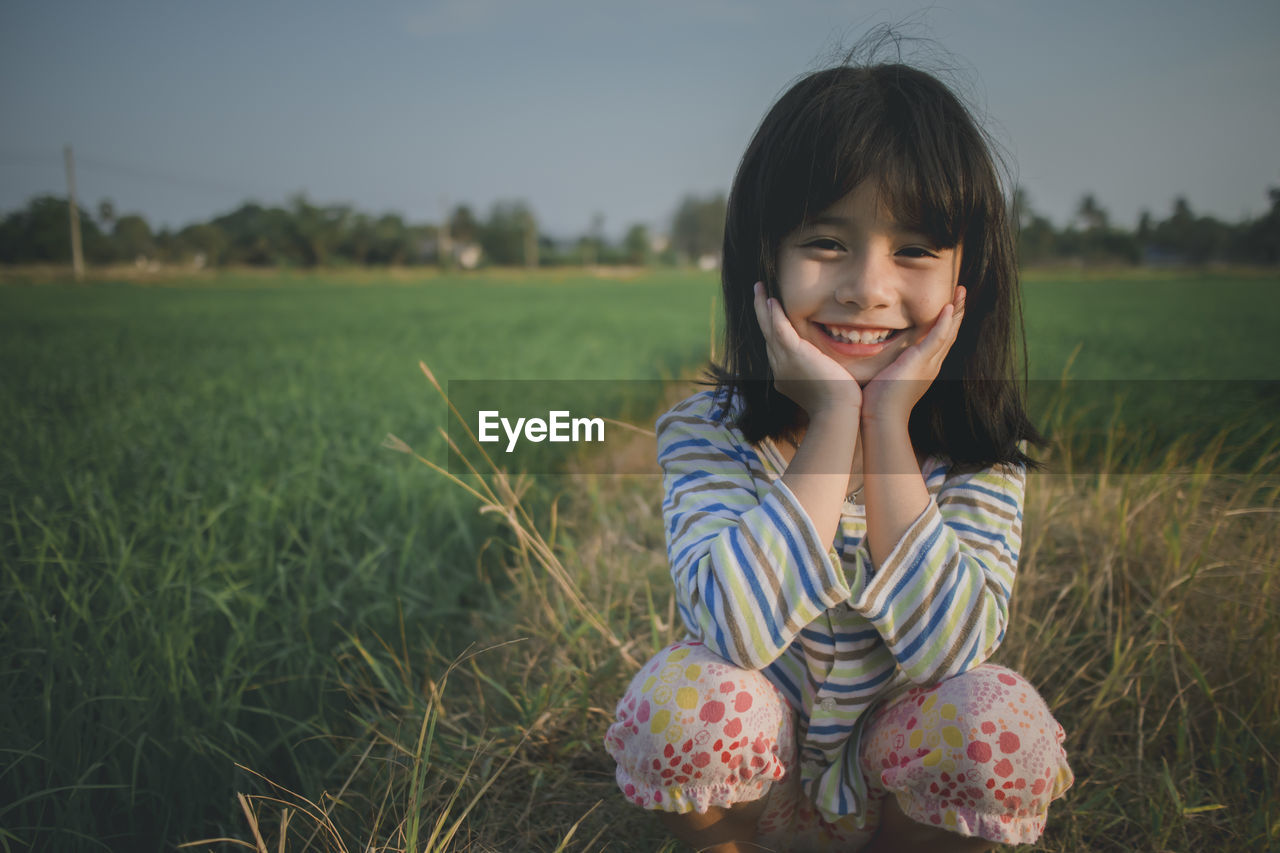 Portrait of cute girl crouching on grassy field against sky