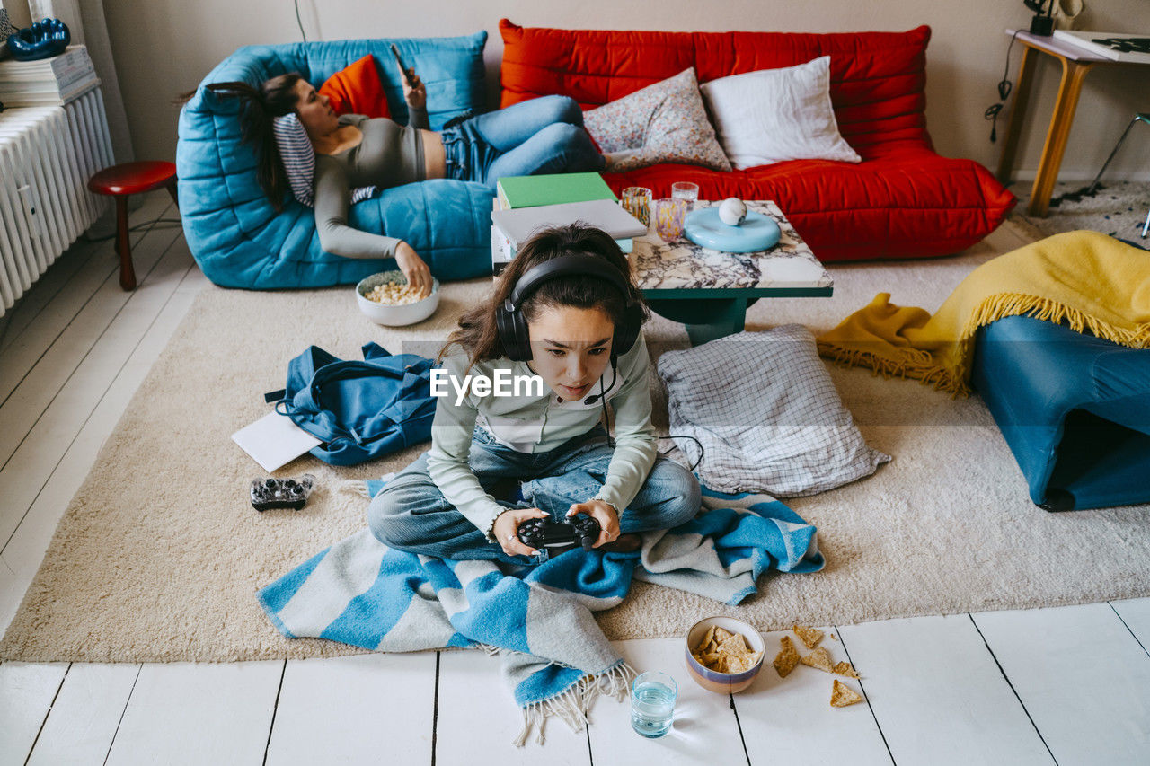 High angle view of woman playing video game while friend using smart phone on sofa at home