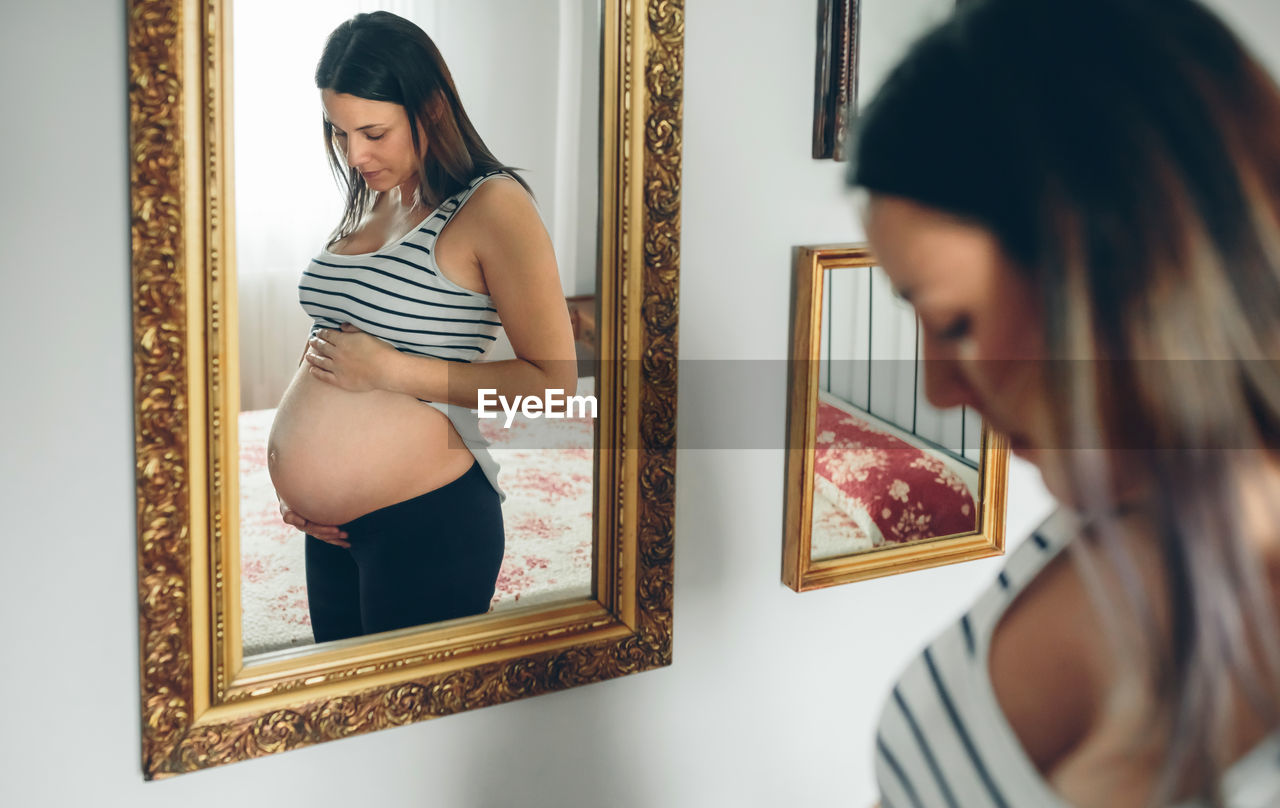 Pregnant woman holding belly while standing by mirror at home