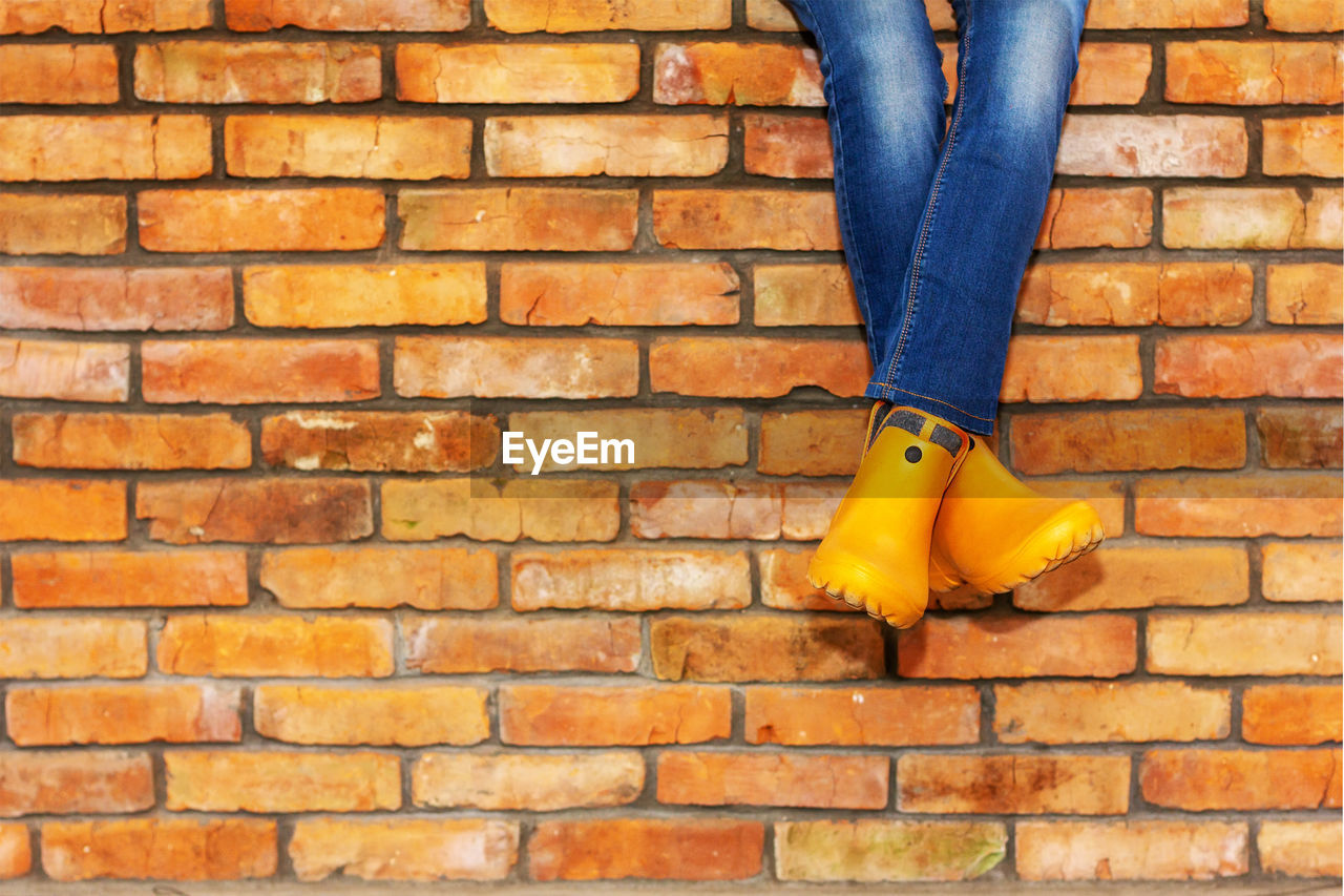 Cross-legged sitting on a brick wall of a young girl in jeans and orange galoshes