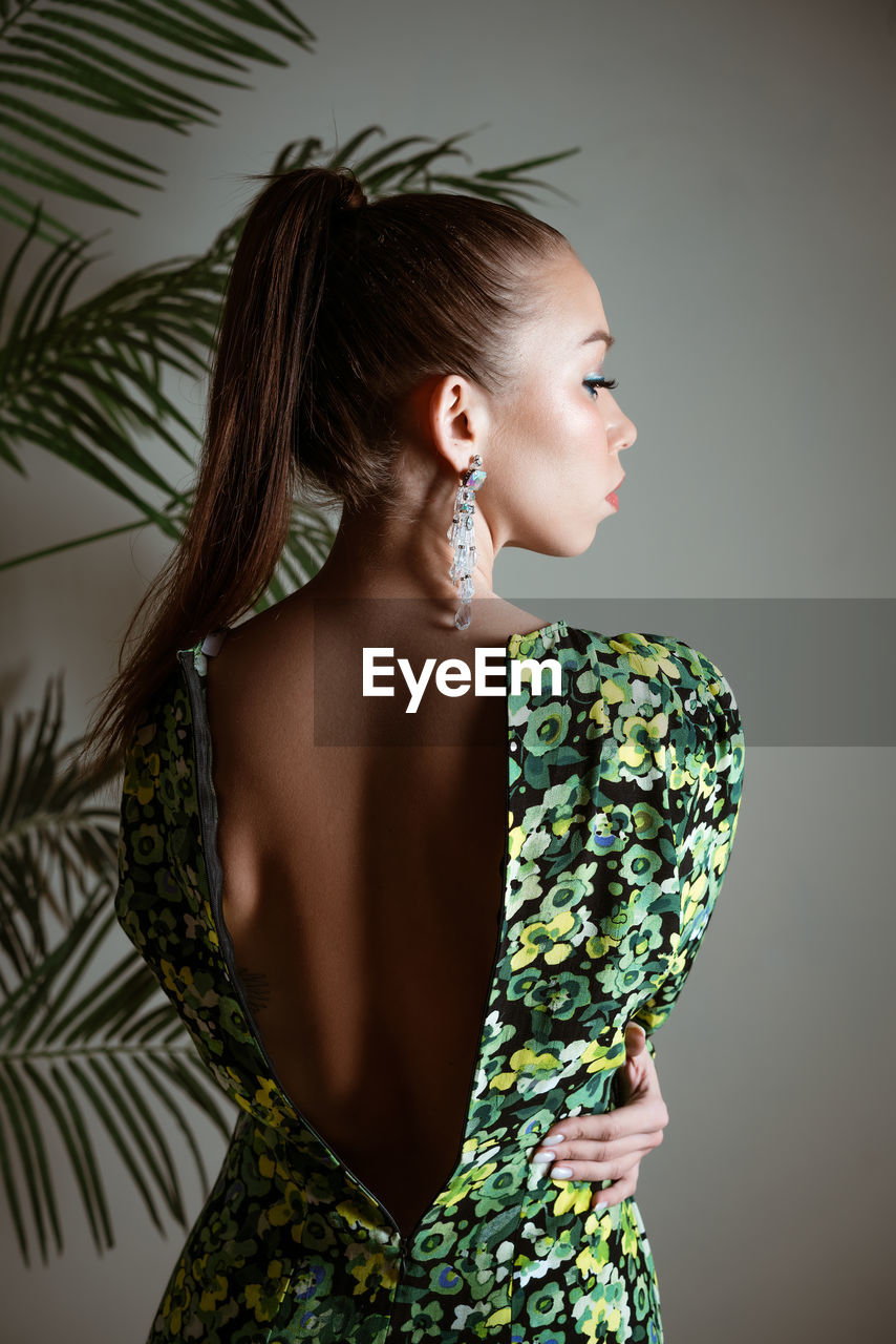 Young woman standing with her back in green floral dress looks to side on gray