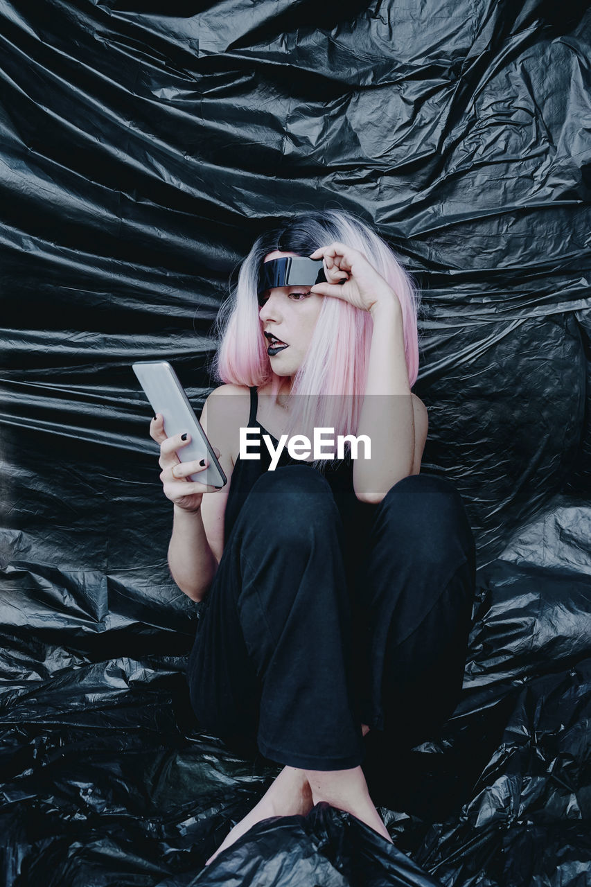 Woman with futuristic eyeglasses using smart phone sitting on black plastic backdrop