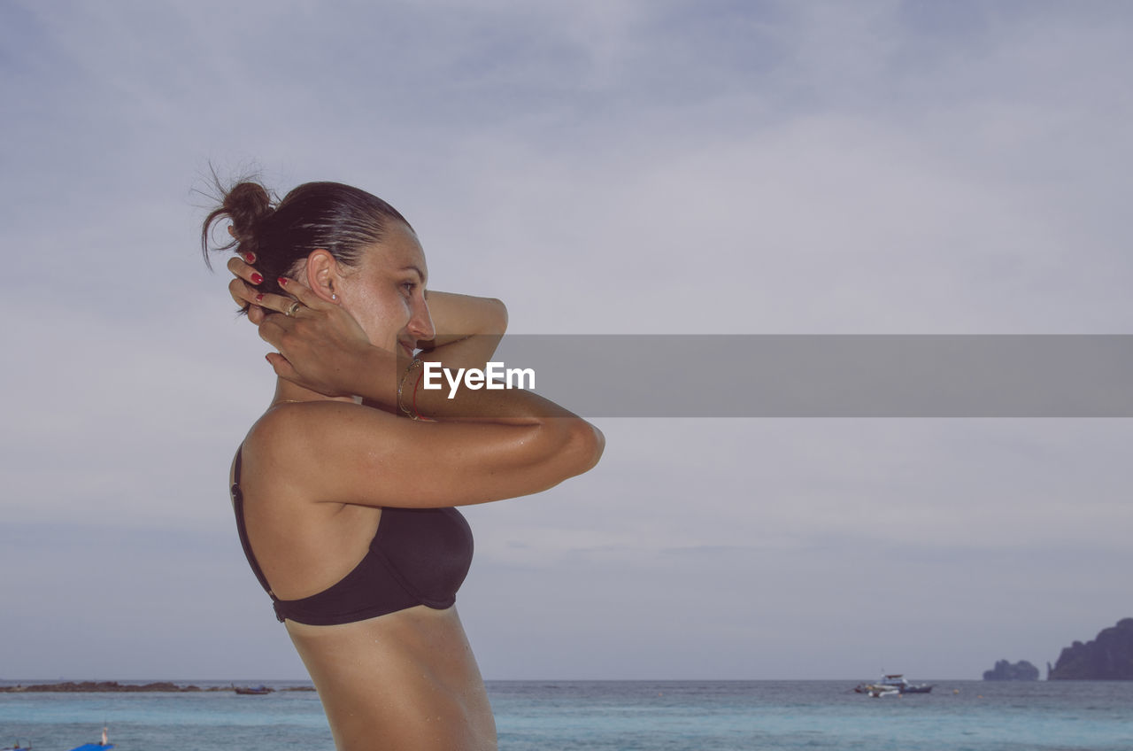 Side view of woman wearing bikini standing by sea against sky