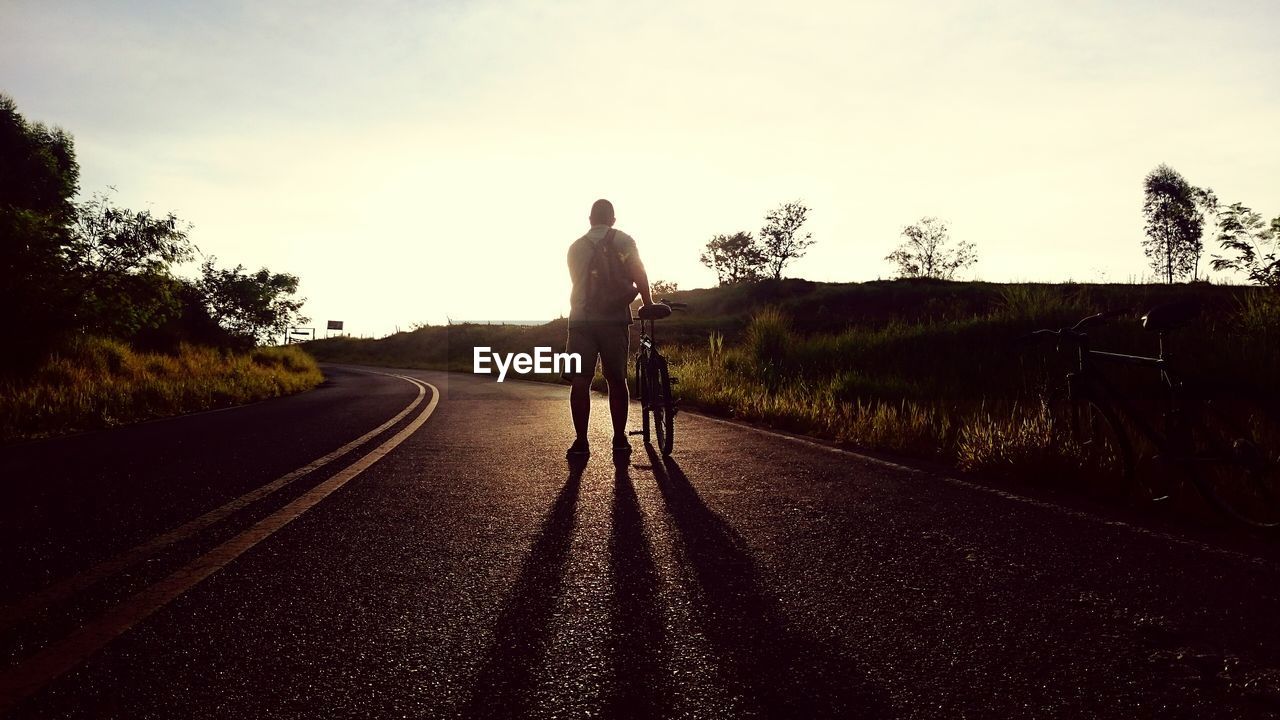 Rear view of man walking with bicycle on country road against sky