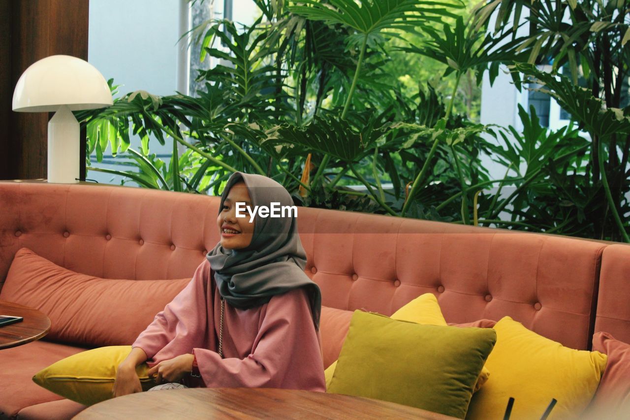 Smiling young woman wearing hijab looking away while sitting on sofa