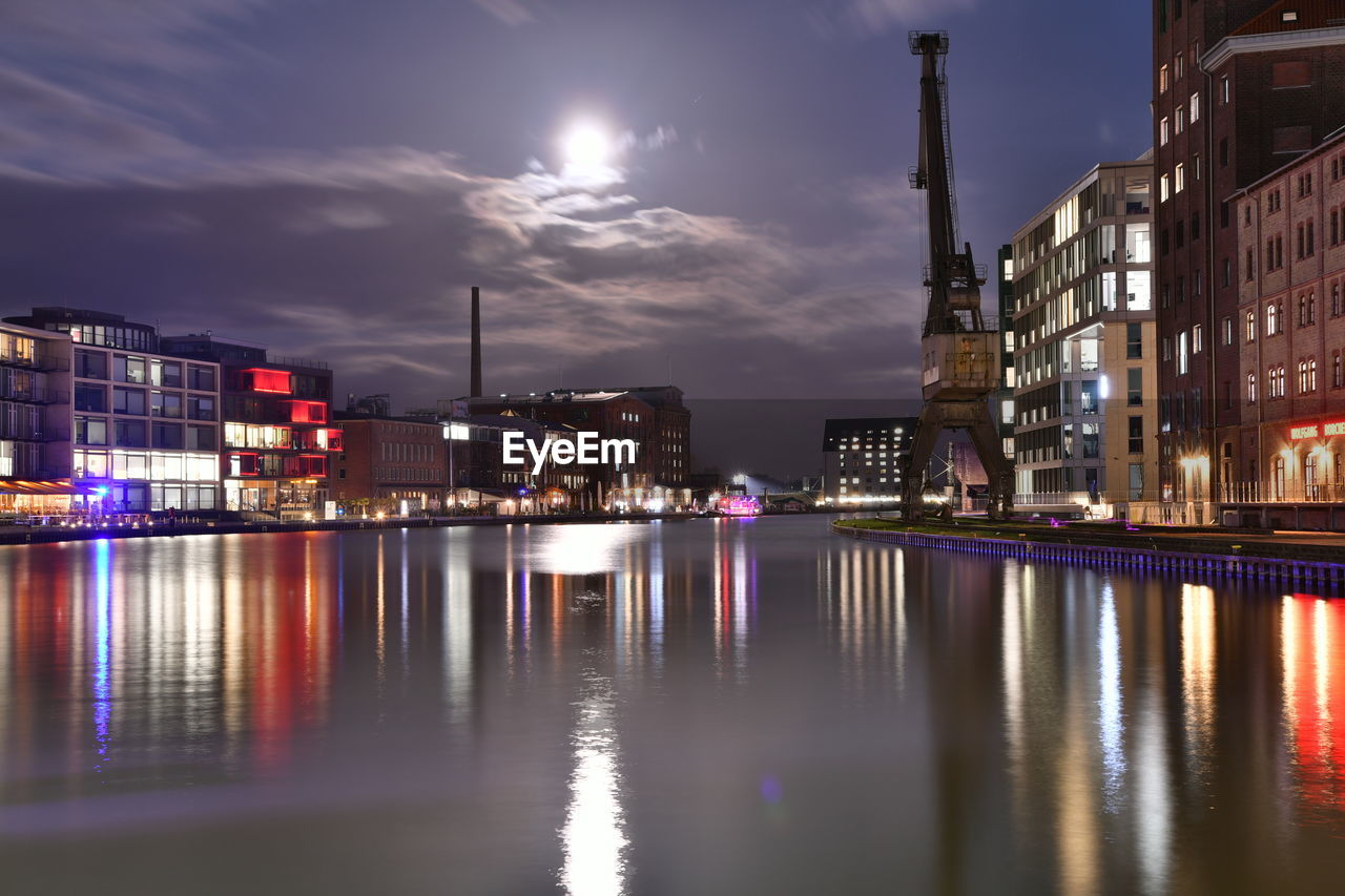 Illuminated buildings by river against sky at night