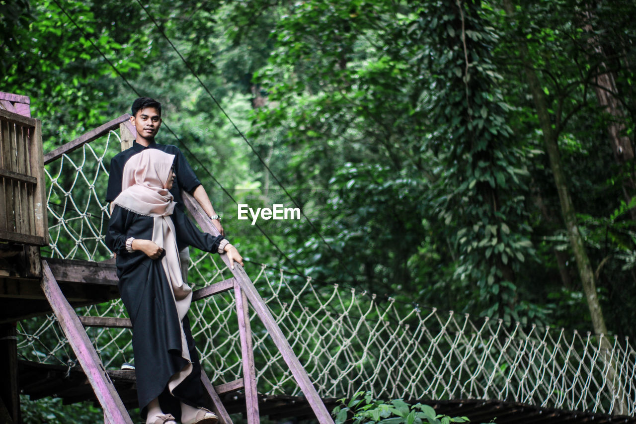 Man and woman on footbridge against trees in forest