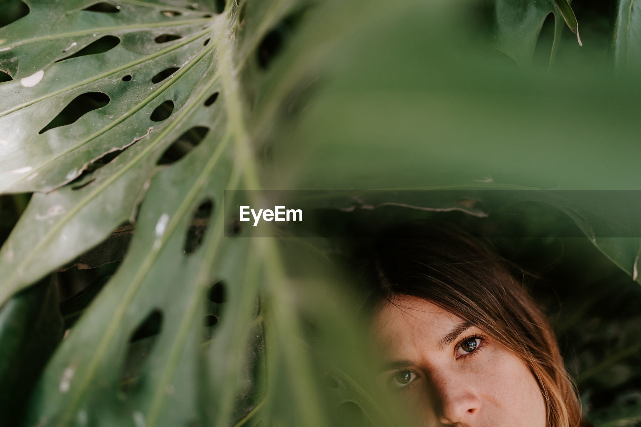 Young serene female standing in tropical park with monstera plants and looking at camera