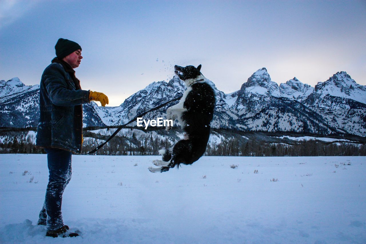 Side view of man playing with dog by mountains on snowy field