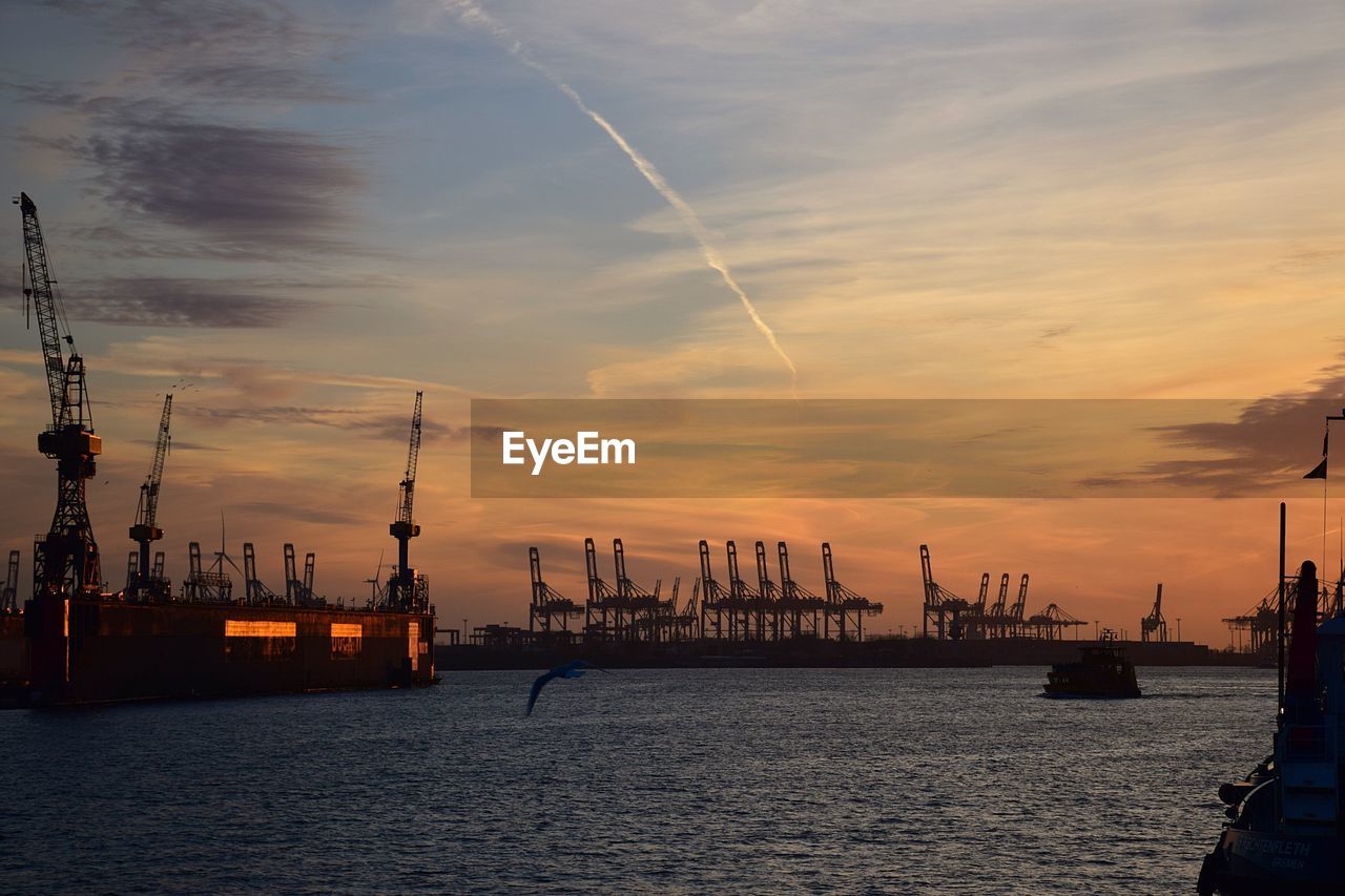 Cranes at commercial dock against sky during sunset