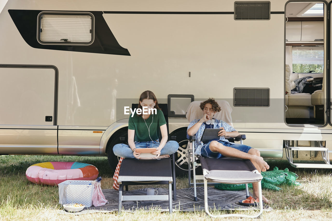 Brother and sister using digital tablet while sitting on folding chairs against motor home