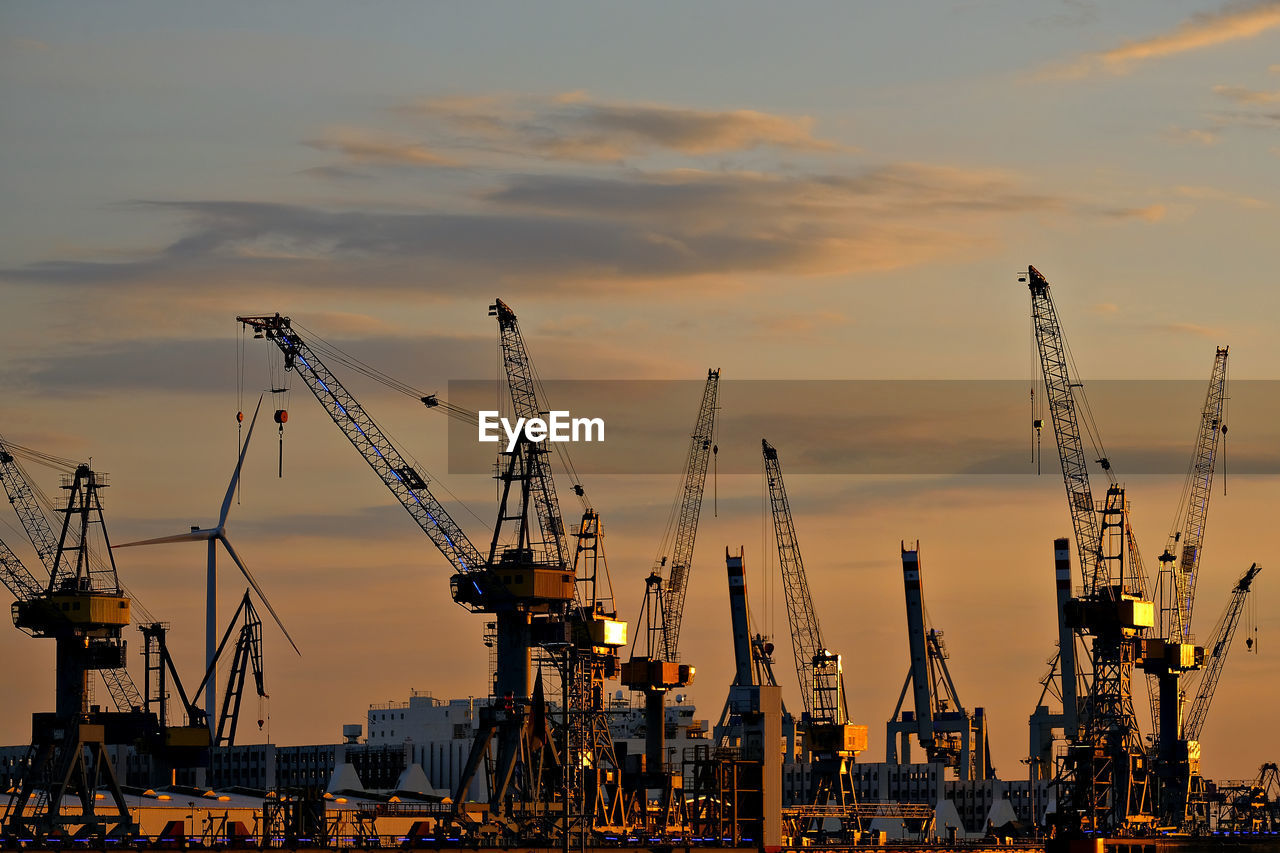 Cranes at harbor against sky during sunset