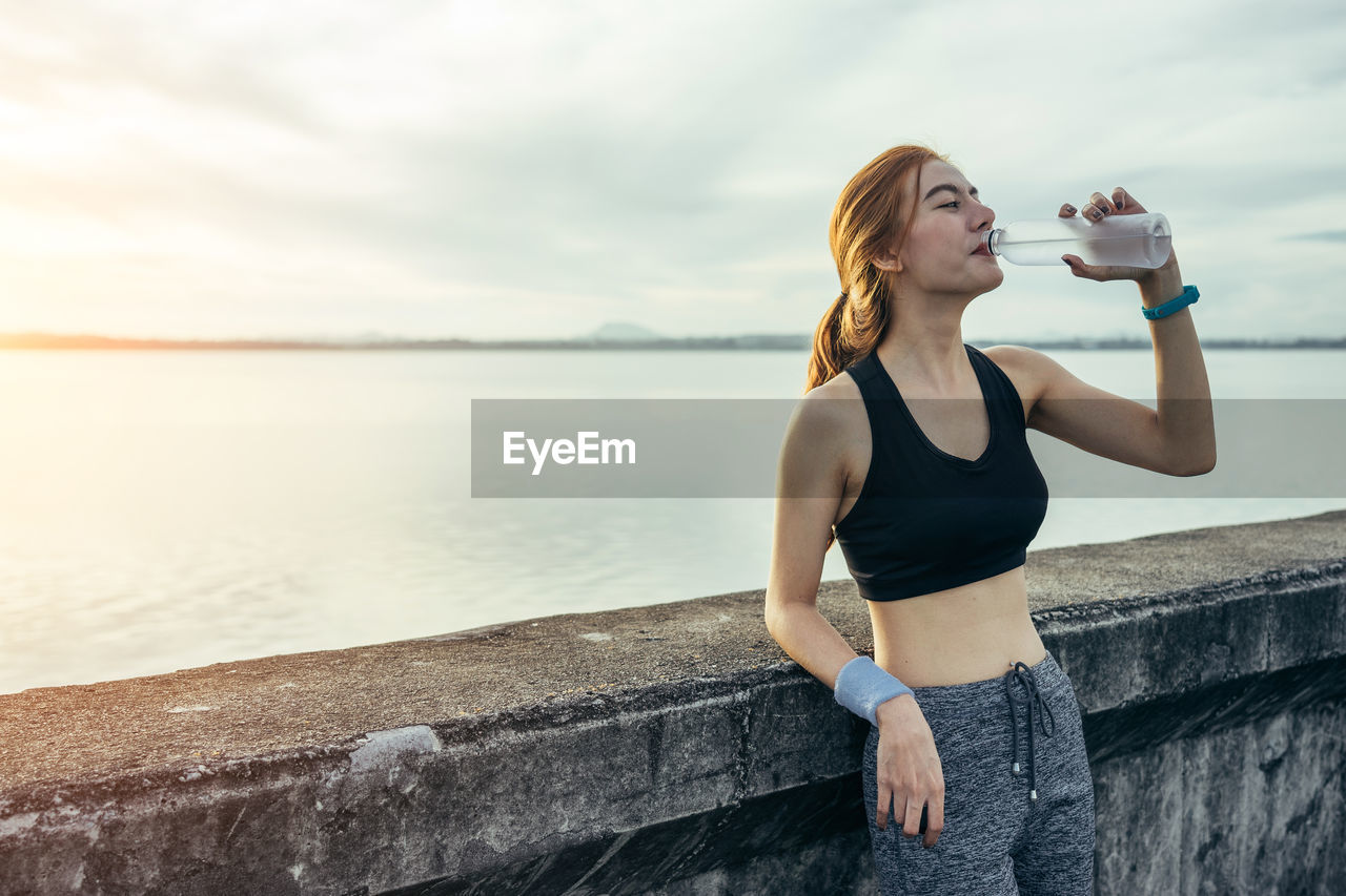 Athlete woman drinking water from bottle while standing by lake against sky