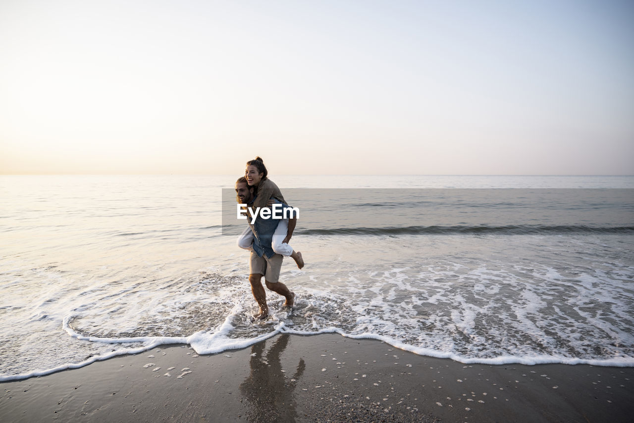 Cheerful man giving piggyback to girlfriend while walking on shore at beach against clear sky during sunset