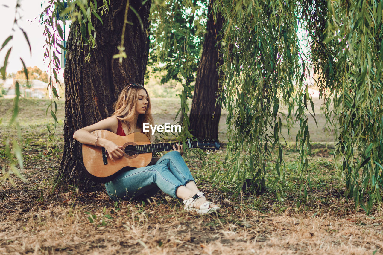 Full length of woman playing guitar while sitting under tree