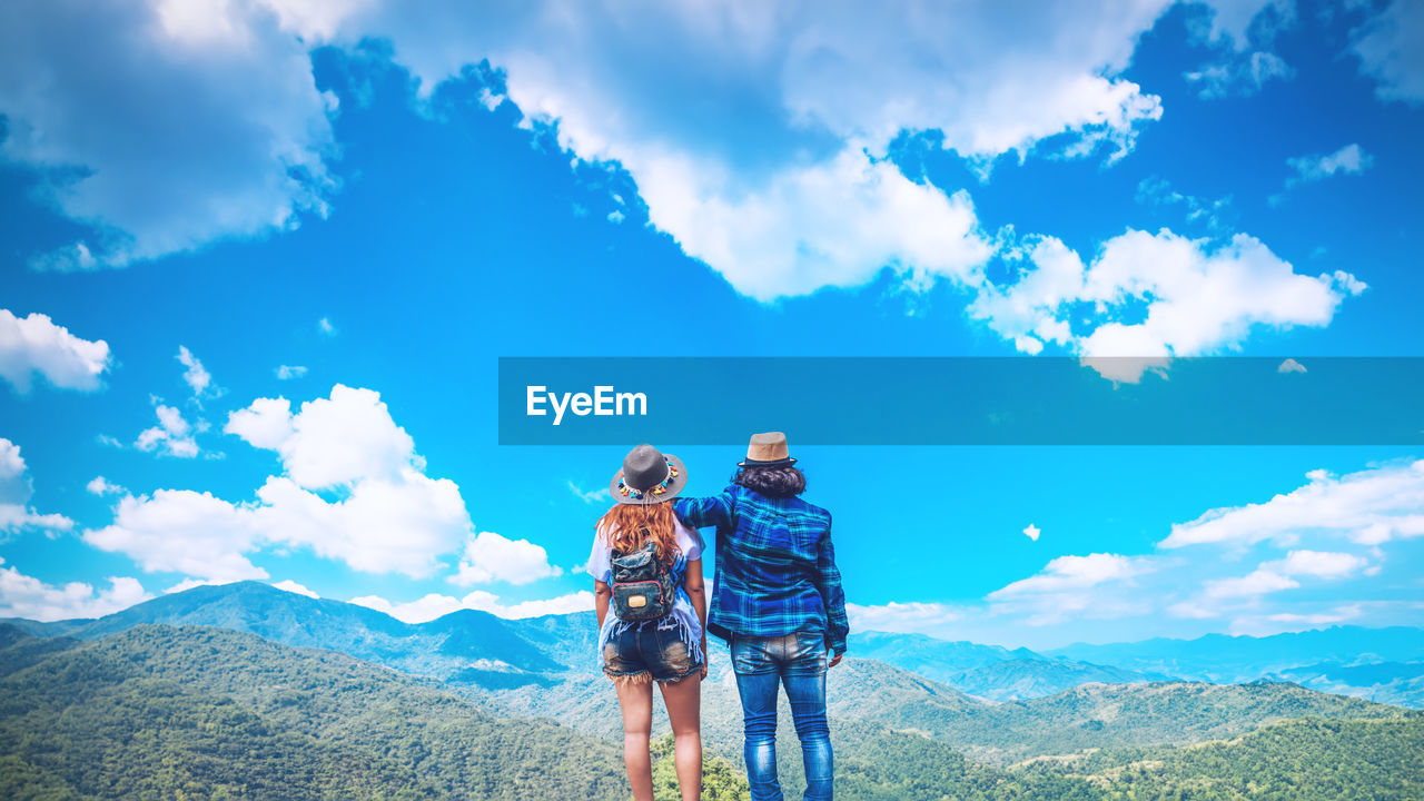 sky, mountain, cloud, adult, two people, activity, hiking, beauty in nature, landscape, nature, scenics - nature, leisure activity, mountain range, environment, men, adventure, togetherness, travel, women, vacation, holiday, trip, travel destinations, emotion, blue, sports, happiness, lifestyles, young adult, backpack, positive emotion, copy space, exploration, outdoors, mountain peak, person, sunlight, female, day, non-urban scene, tourism, full length, friendship, land, recreation, tourist, smiling, tranquil scene, journey, bonding, looking at view, idyllic, summer, relaxation, tranquility, love, standing, forest
