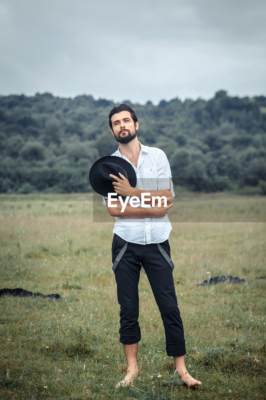 Young man holding hat while standing on grassy field against cloudy sky