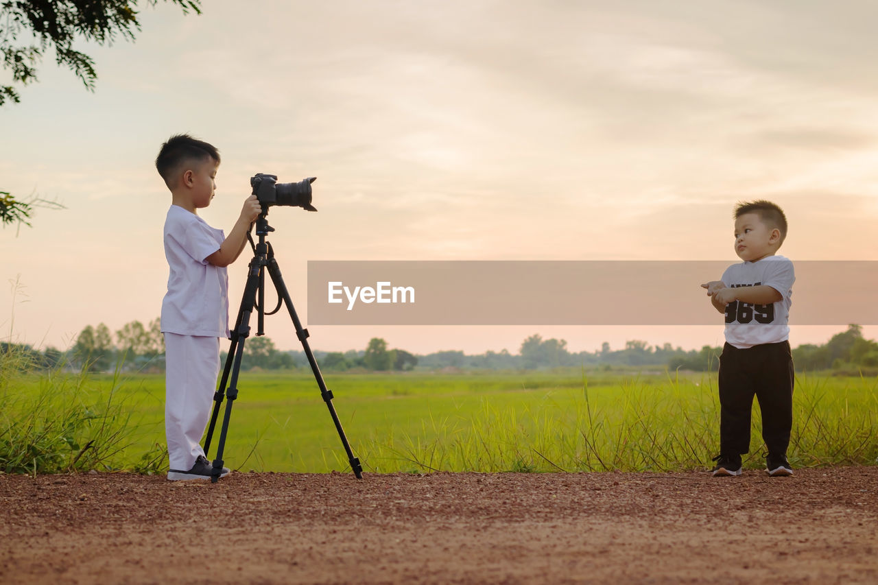 Boy photographing cute brother on land against sky during sunset