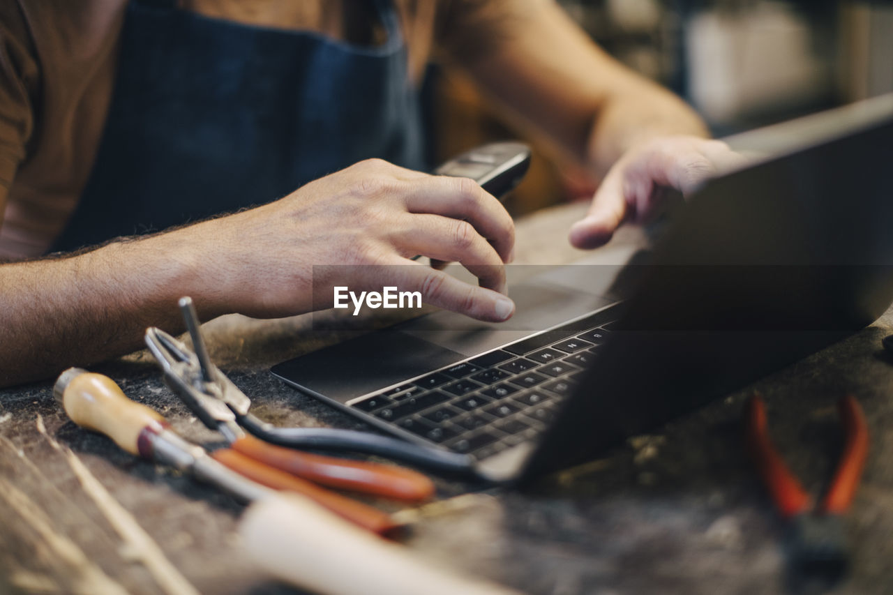 Midsection of upholstery worker using laptop while sitting at workbench in workshop