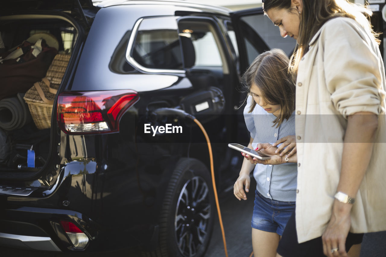 Portrait of mother, father and two daughters standing by car at electric vehicle charging station