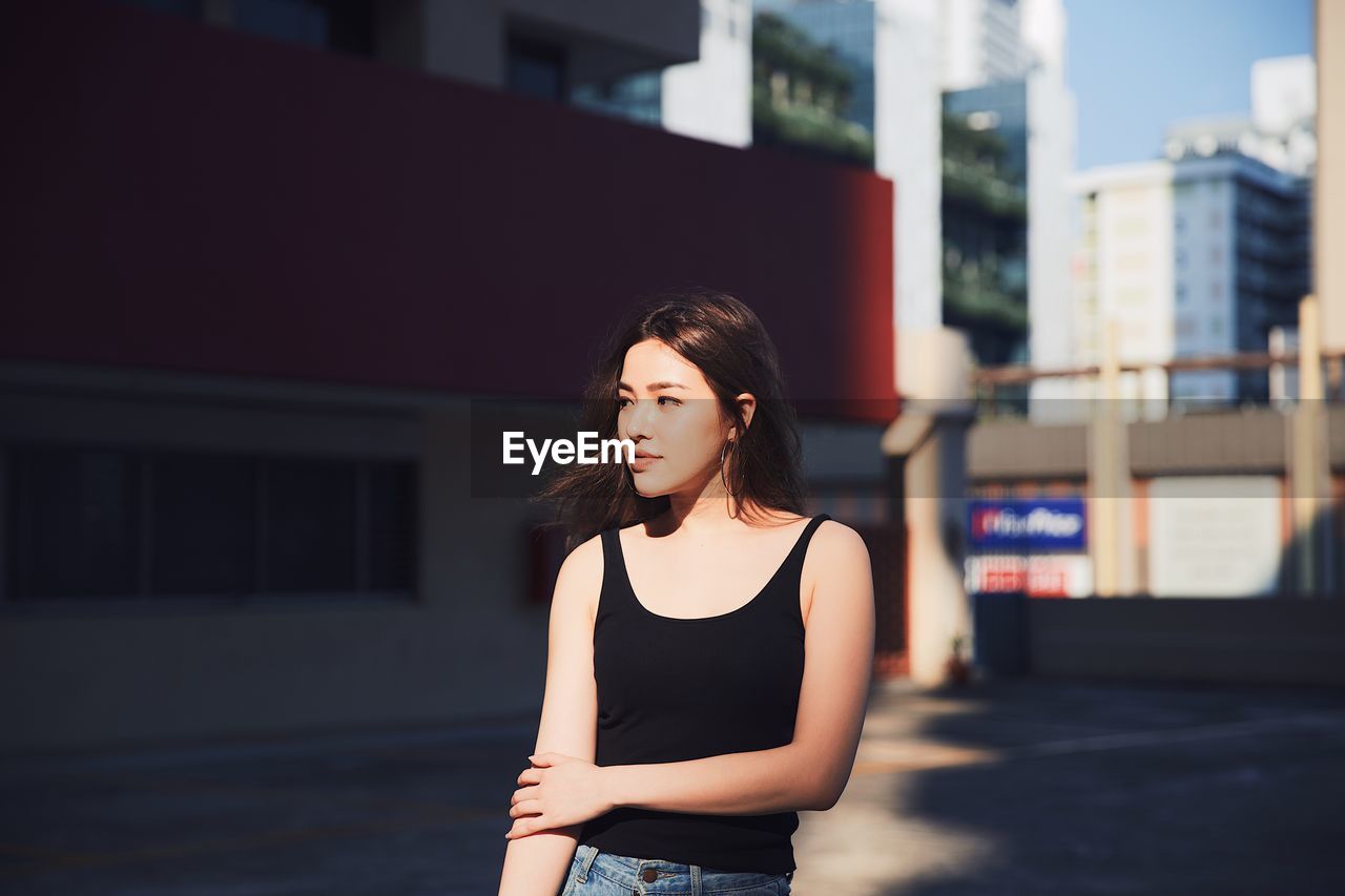 Young woman looking away while standing on road against buildings in city