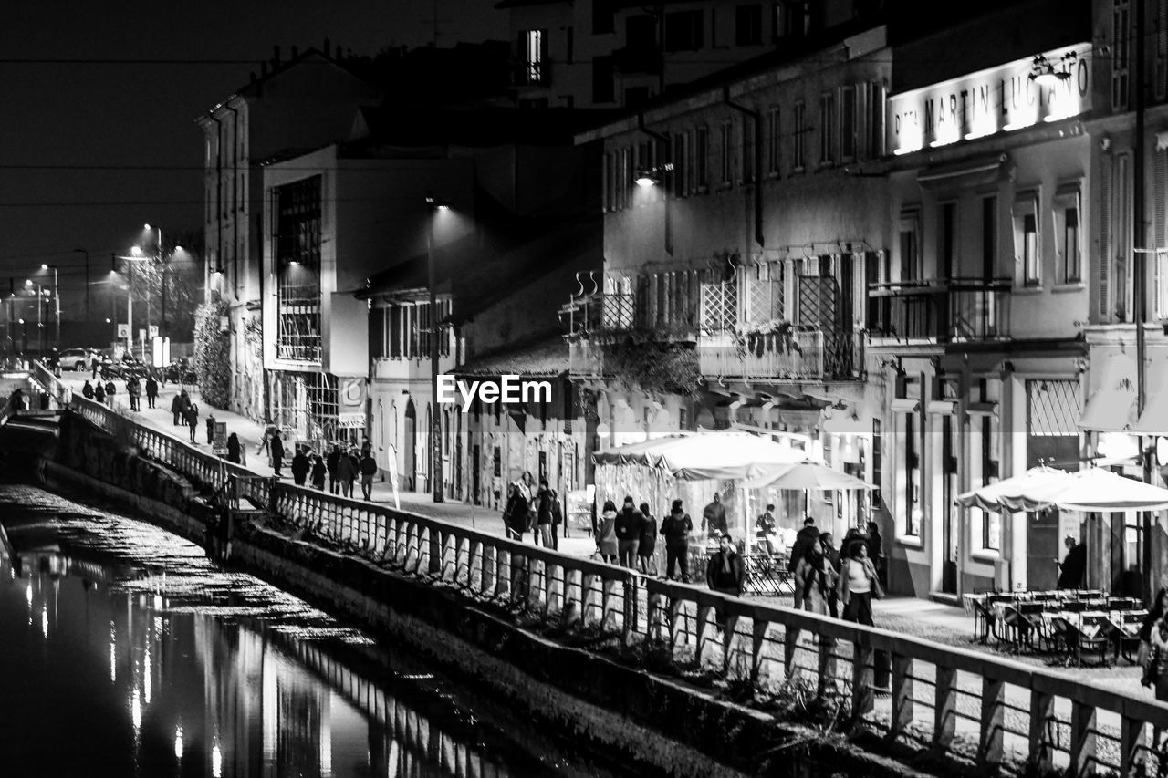 PEOPLE ON ILLUMINATED STREET AMIDST BUILDINGS IN CITY