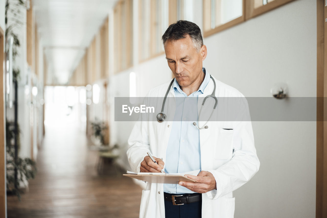 Mature doctor writing medical record at corridor in hospital