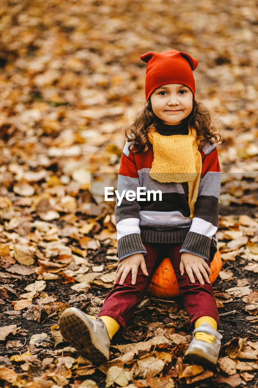 Happy cute little girl child in warm bright clothes sit on a pumpkin in the autumn forest outdoors