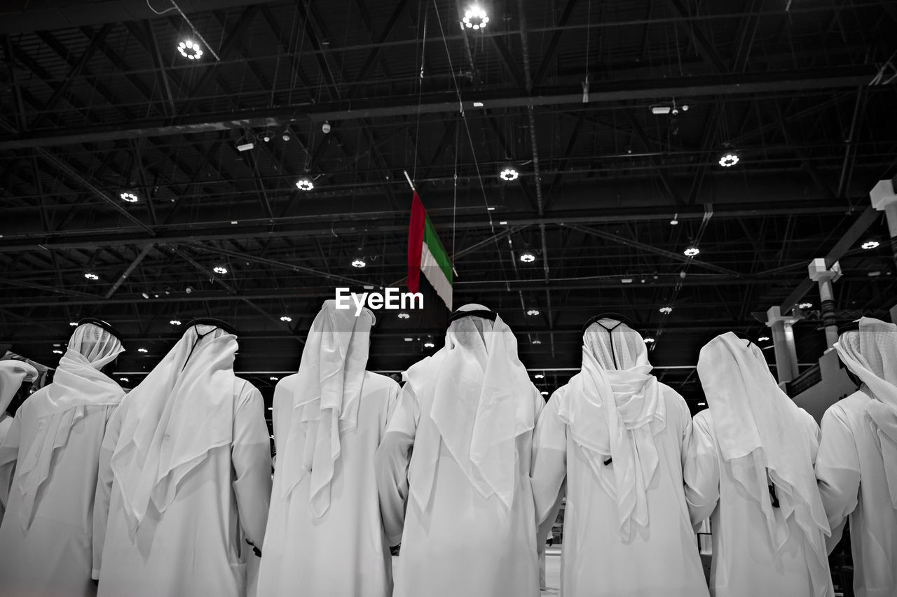 Rear view of men wearing white traditional clothing with united arab emirates flag hanging from ceiling in background