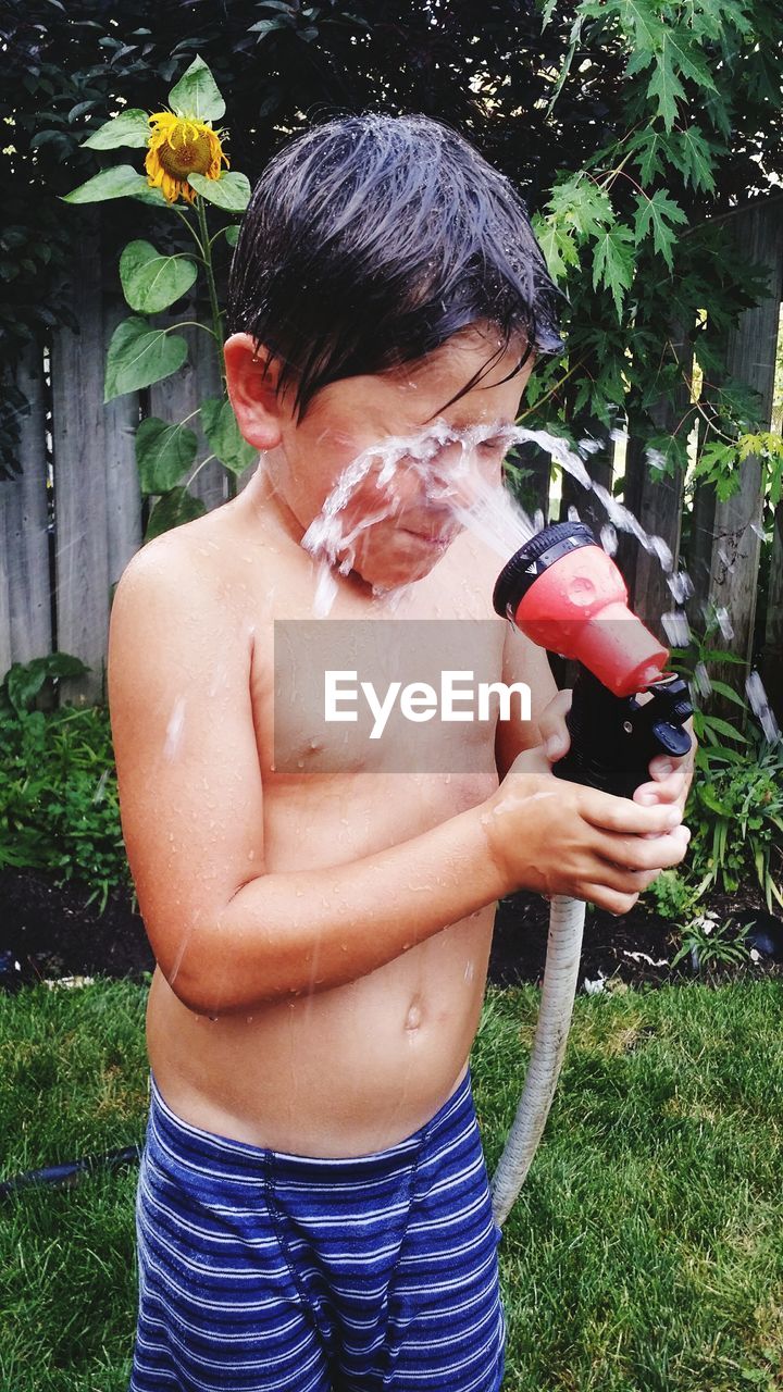 Shirtless boy splashing water with hose while standing in backyard