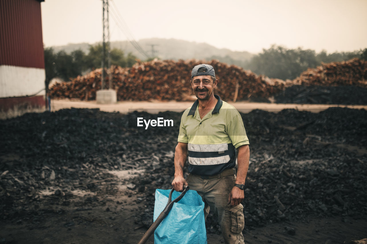 Smiling worker with plastic bag and tool standing at coal factory