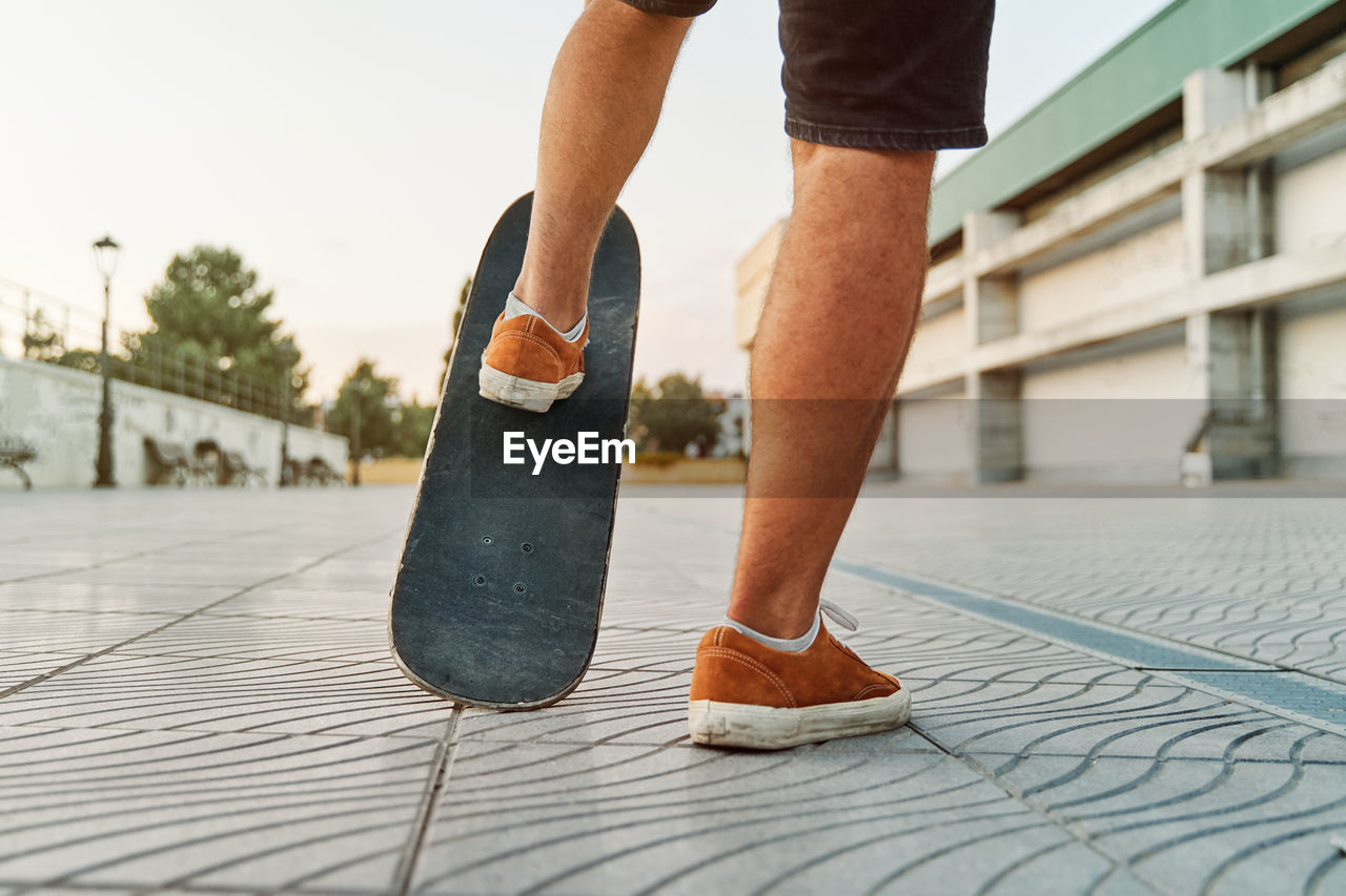 Unrecognizable talented male in sneakers riding shabby skateboard along street at sunset in summer