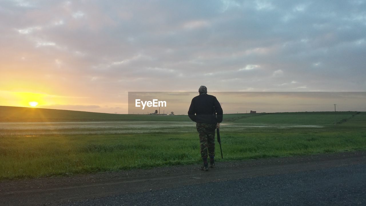 Rear view of military man with rifle walking on field against cloudy sky