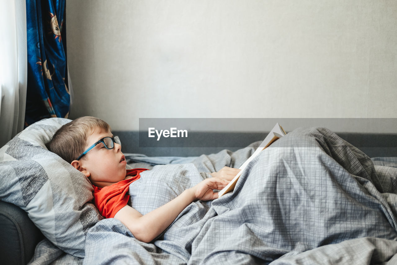 Boy with glasses lies on a bed under a blanket and reads a book. 