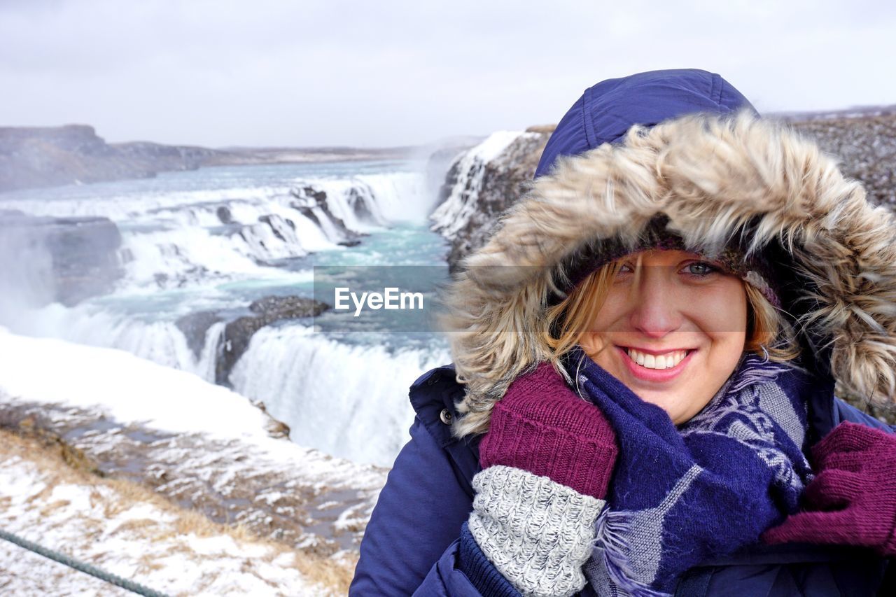 Close-up portrait of smiling young woman standing against waterfall