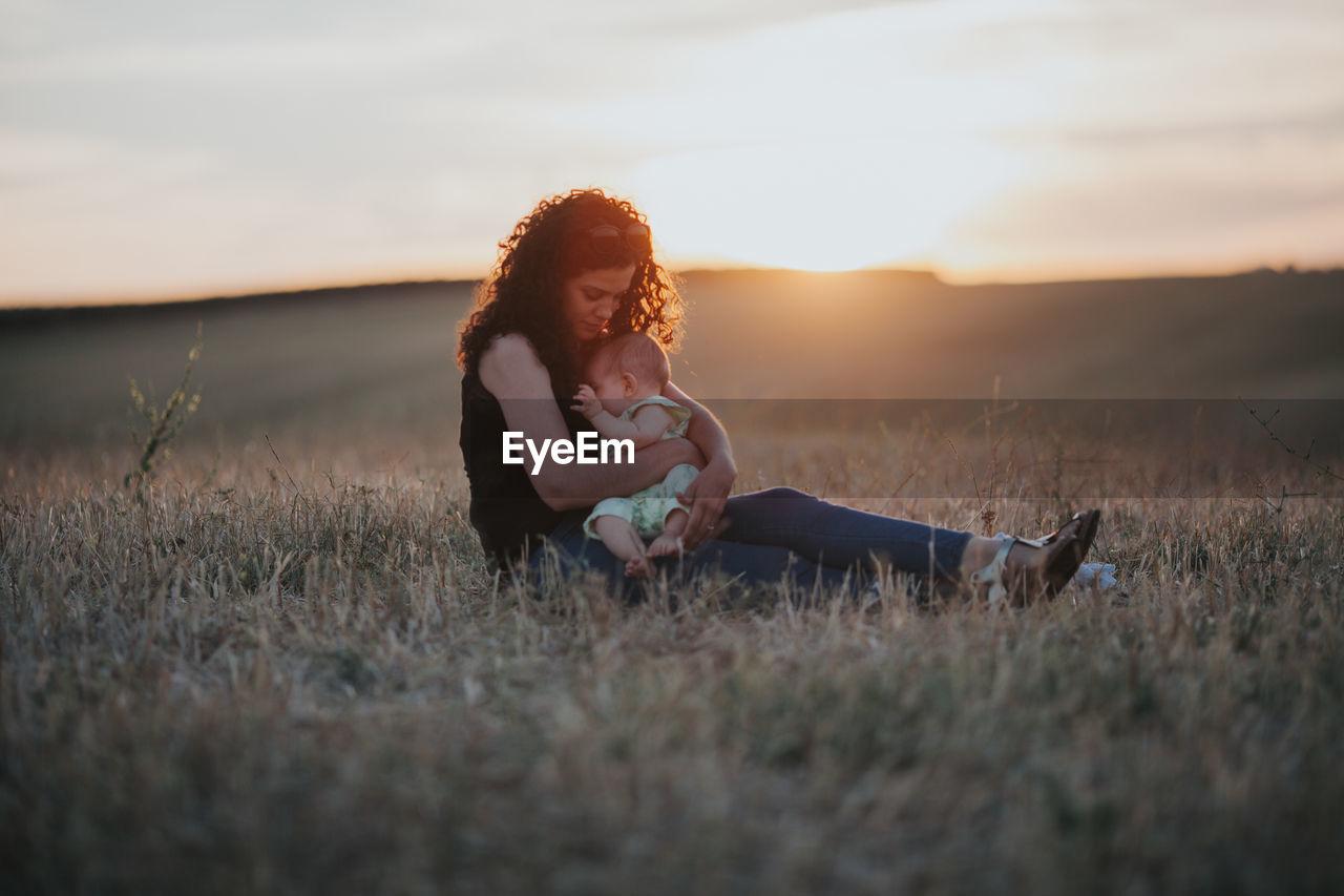 Woman with daughter sitting on grass against sky during sunset