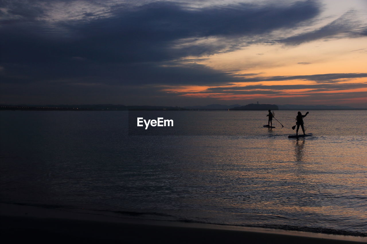 Silhouette people paddleboarding in sea against sky during sunset