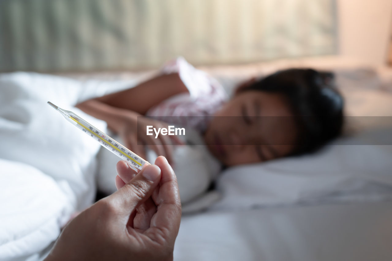 Cropped hand of mother holding thermometer while daughter sleeping on bed