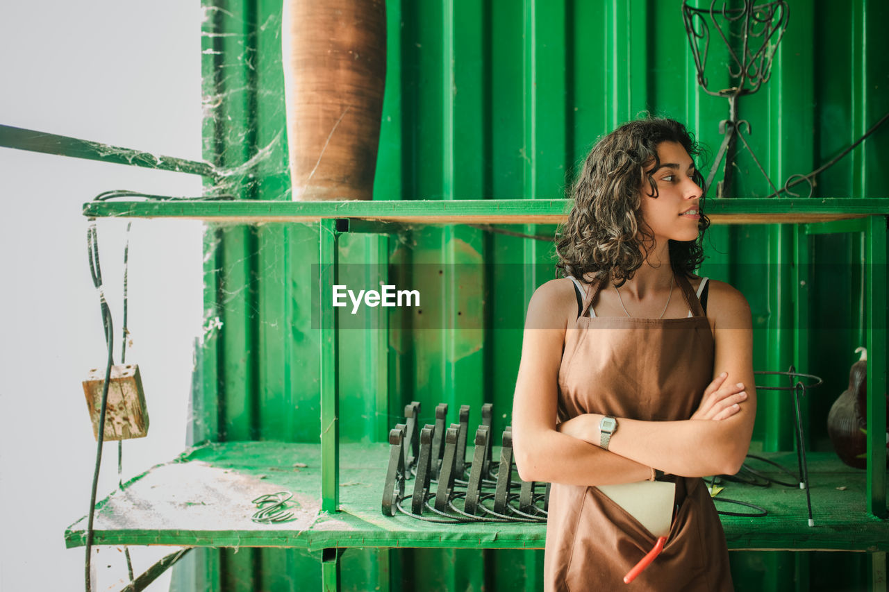 Beautiful young woman standing against green barrel at pottery shop