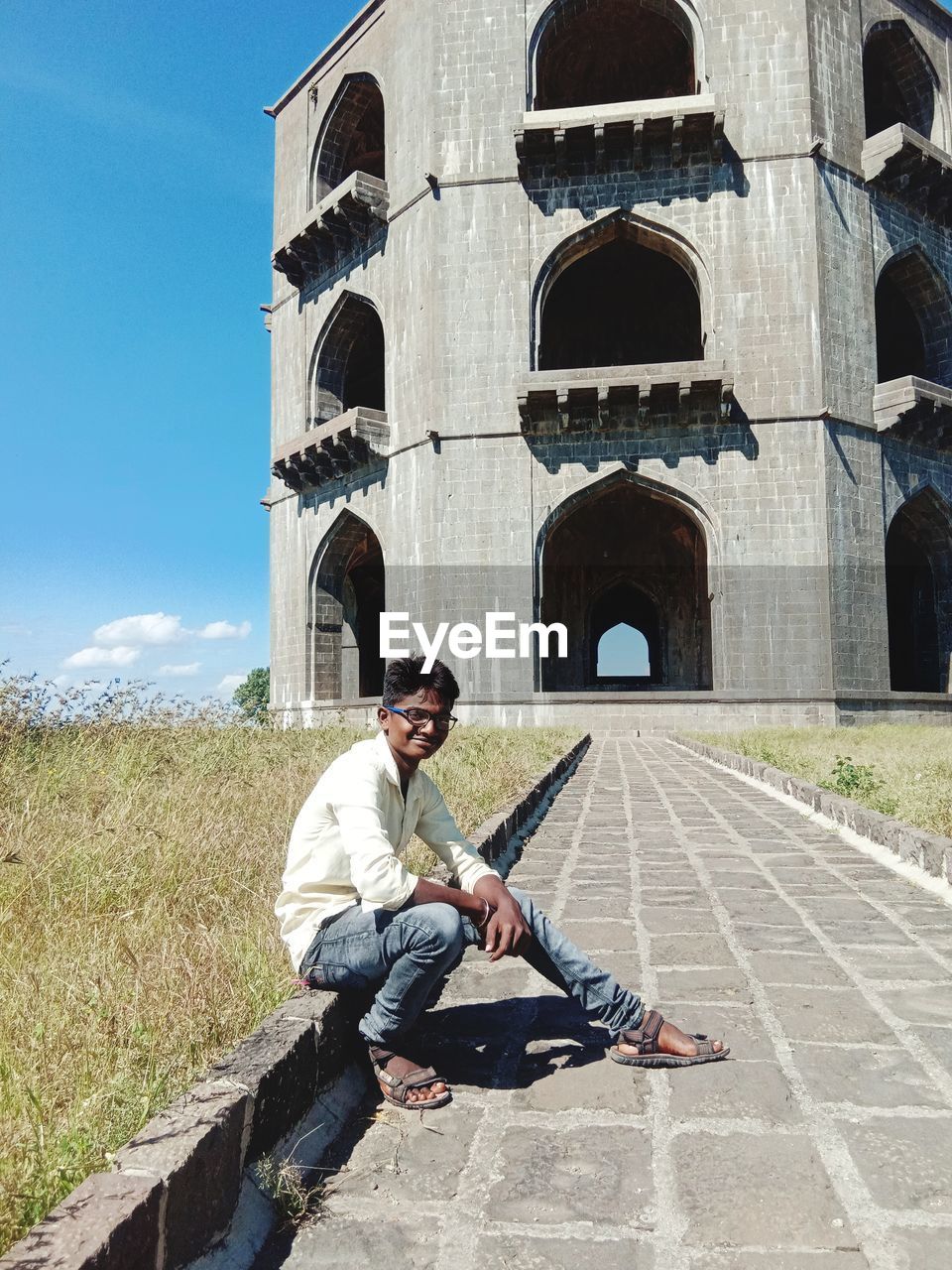 Full length of man sitting on footpath against building and sky