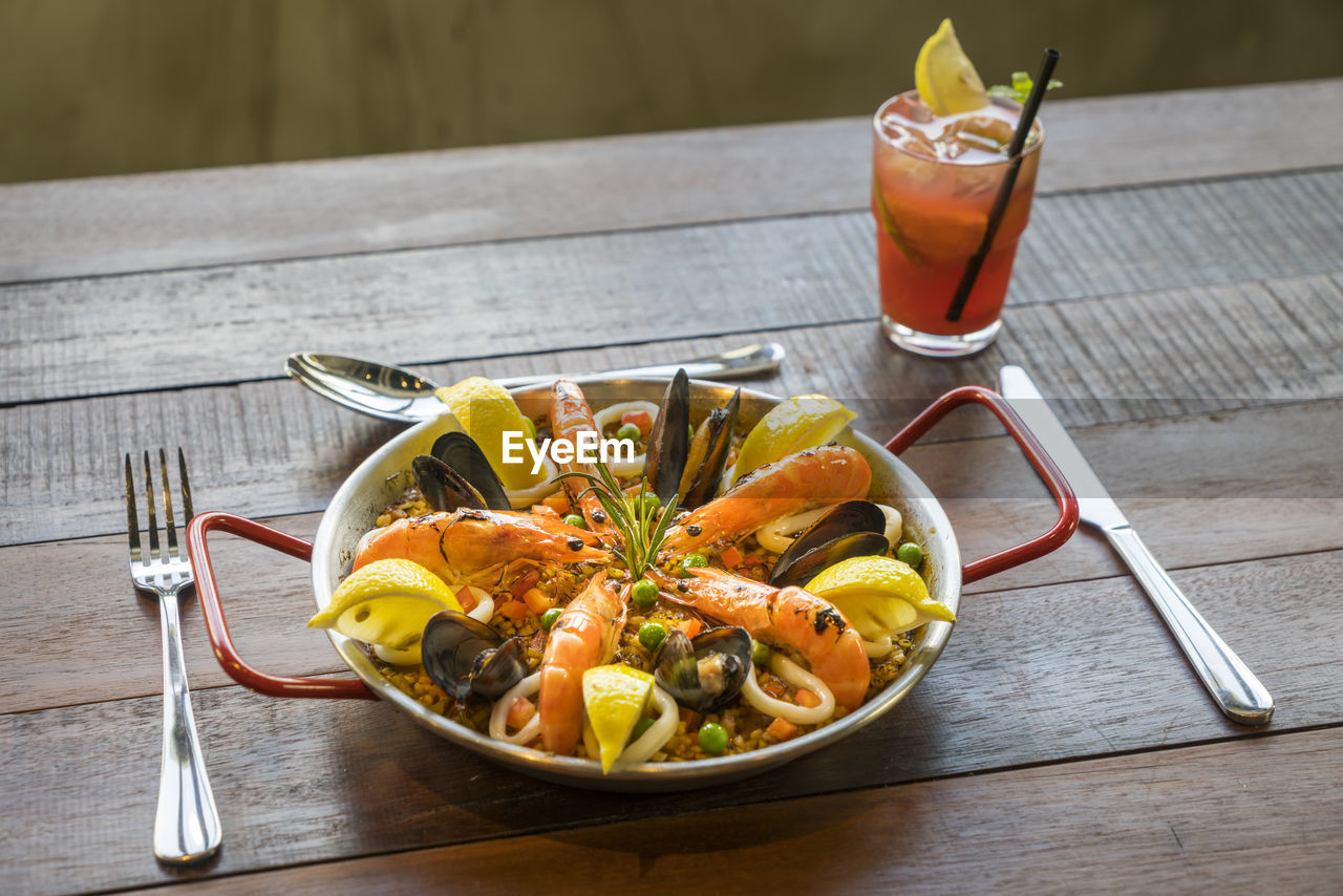 High angle view of paella served in bowl with drink on wooden table