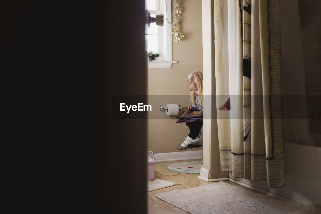 Girl using tablet computer while sitting in bathroom seen through doorway