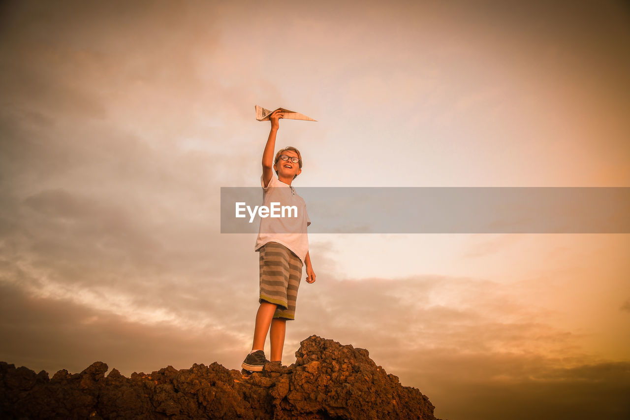 Low angle view of boy holding paper airplane against sky during sunset