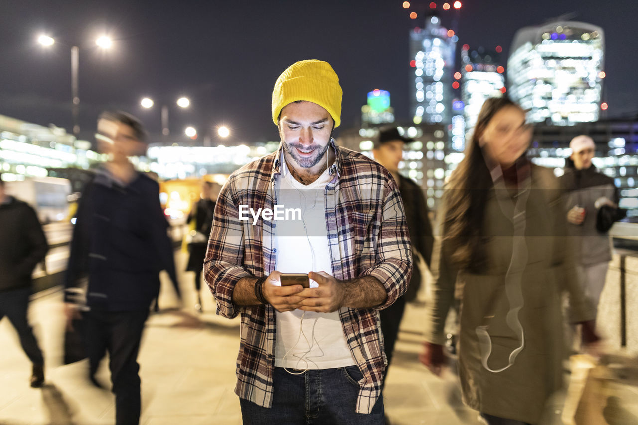 Uk, london, smiling man looking at his phone by night with blurred people passing nearby