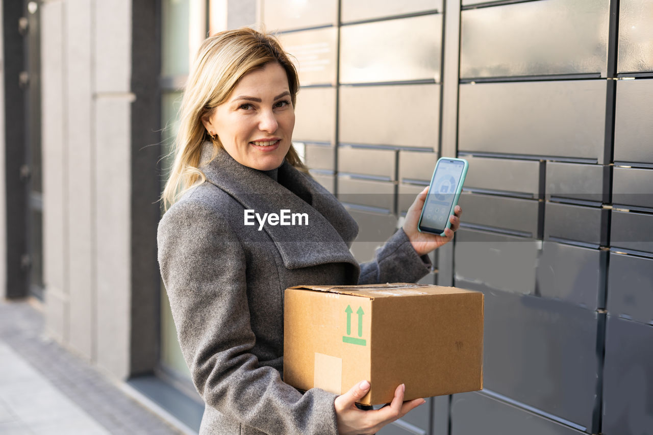Beautiful woman picking up a package from a smart electronic steel parcel locker box, automatic
